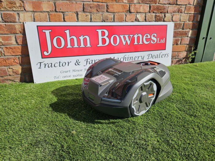 Husqvarna Automower® 410XE NERA robotic lawn mower operating on a well-kept lawn. The mower is shown near the lawn’s edge, highlighting its EdgeCut feature. Surrounded by a neat garden, the compact mower demonstrates its smart design, built-in sensors for obstacle avoidance, and suitability for lawns up to 1000 m².