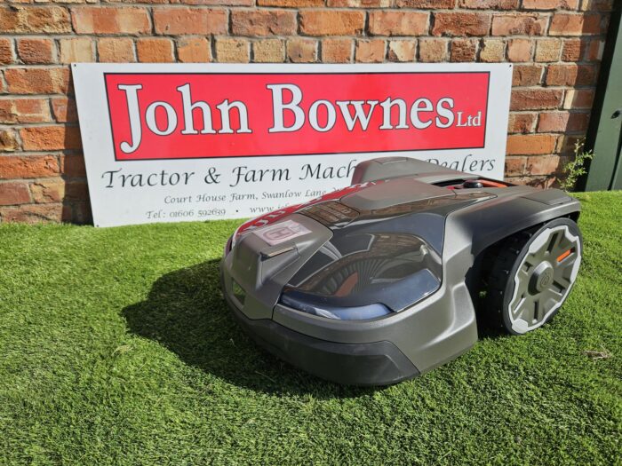 Husqvarna Automower® 430X NERA robotic lawn mower operating on a large, uneven lawn. The mower is shown navigating rugged terrain near garden plants, demonstrating its robust design, obstacle detection sensors, and wire-free satellite installation capability. Ideal for lawns up to 3200 m².