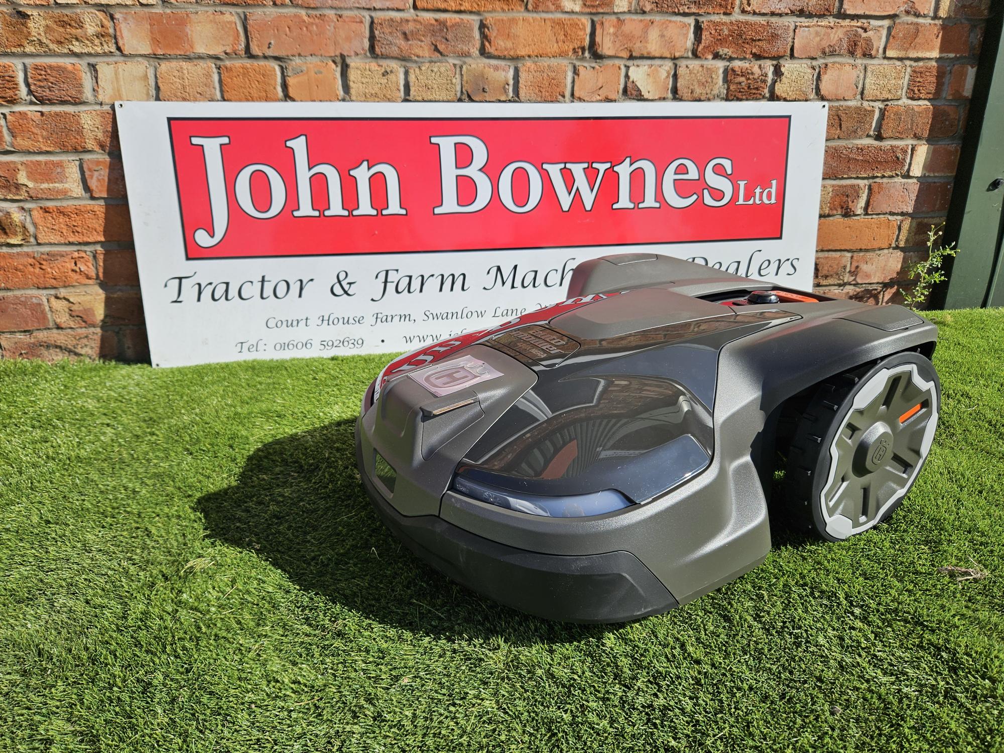 Husqvarna Automower® 430X NERA robotic lawn mower operating on a large, uneven lawn. The mower is shown navigating rugged terrain near garden plants, demonstrating its robust design, obstacle detection sensors, and wire-free satellite installation capability. Ideal for lawns up to 3200 m².